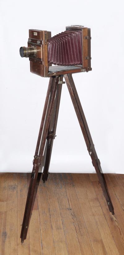Фотокамера со штативом. Одна из ранних студийных фотокамер. На объективе ещё отсутствует механический затвор с выдержкой съемки.